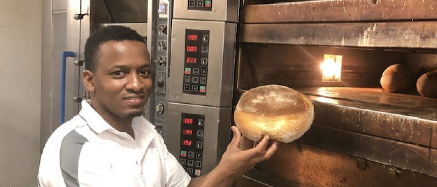 Junger Mann mit einem Brot in der Hand steht vor großem Bäckerofen