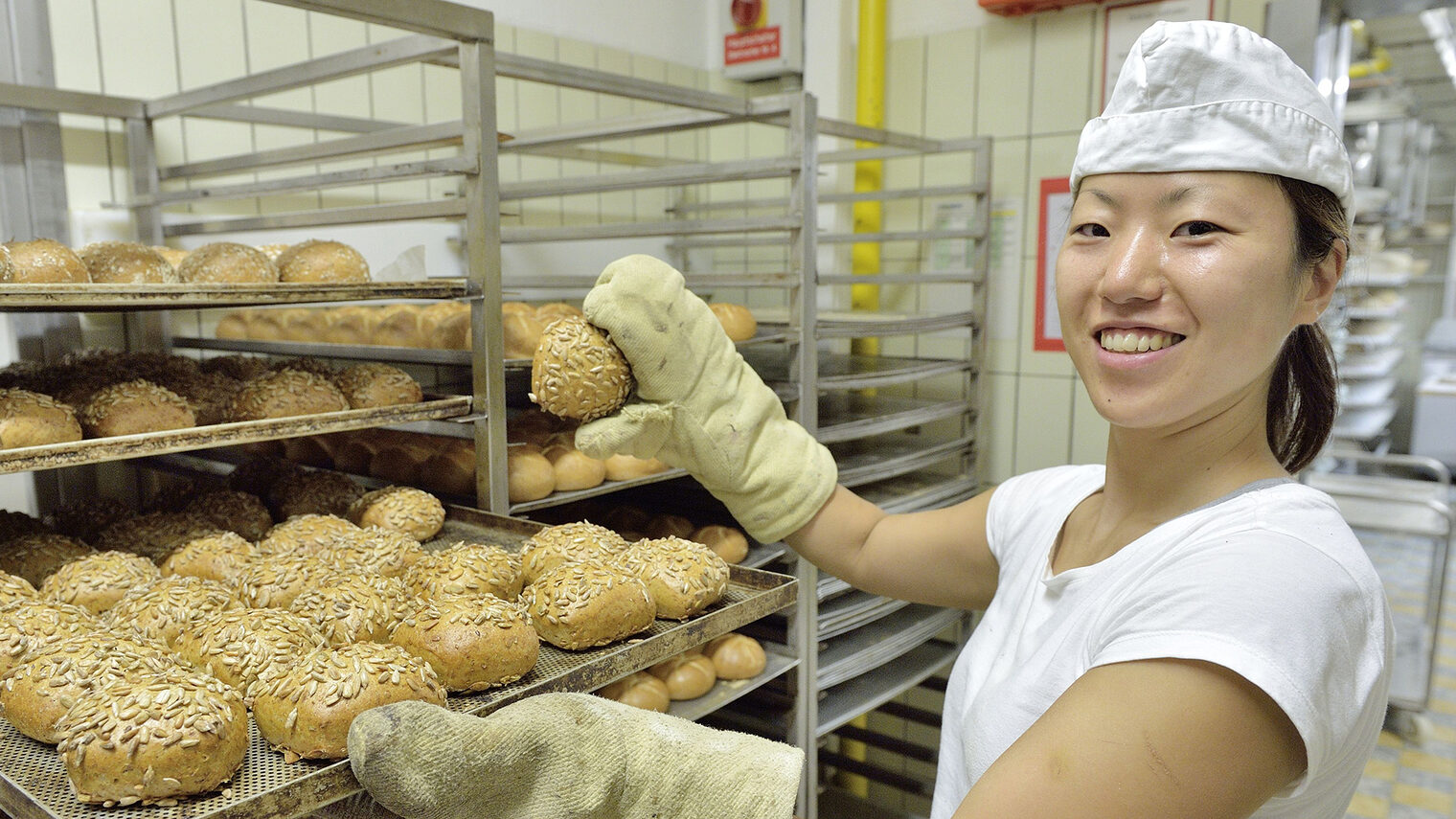 Junge Bäckerin mit Backblech voller Brötchen lächelt in die Kamera.
