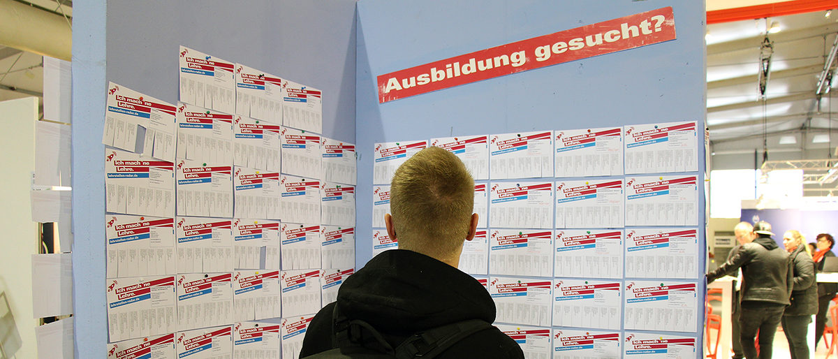 Qualifizierte Mitarbeiter finden Junger Mann steht vor einer Stellwand mit Aufschrift "Ausbildung gesucht?" auf einer Messe