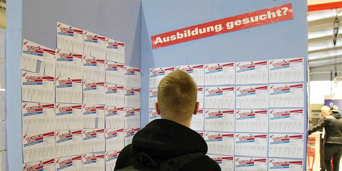 Qualifizierte Mitarbeiter finden Junger Mann steht vor einer Stellwand mit Aufschrift "Ausbildung gesucht?" auf einer Messe