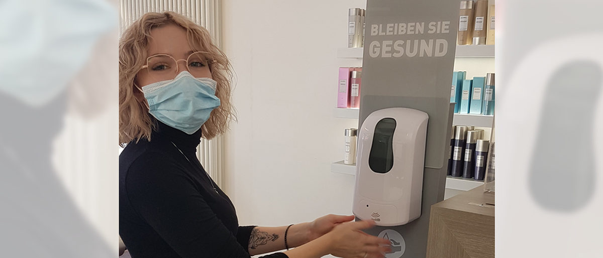Für Julia Harzer ist die Einhaltung der strengen Corona-Auflagen eine Selbstverständlichkeit. Julia Harzer mit Mundschutz betätigt Desinfektionsmittelspender in Friseursalon.