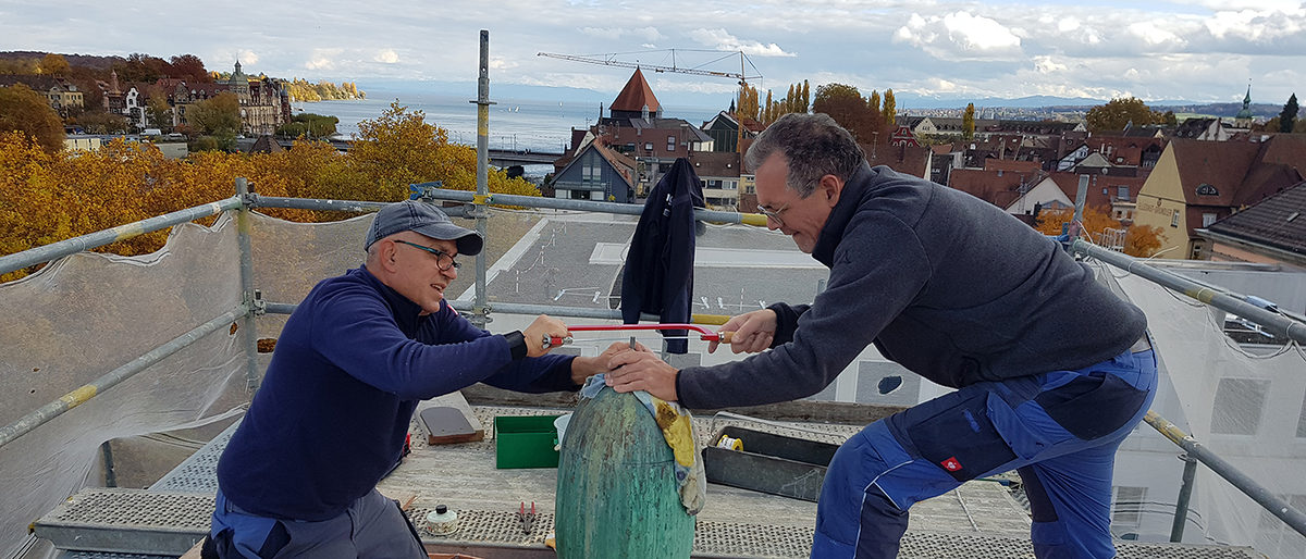 Thomas Pfingst (rechts) und sein Mitarbeiter XY bei der Arbeit. Thomas Pfingst (rechts) bearbeitet mit seinem Mitarbeiter XY die Messing-Spitze auf dem Handwerkskammerdach.