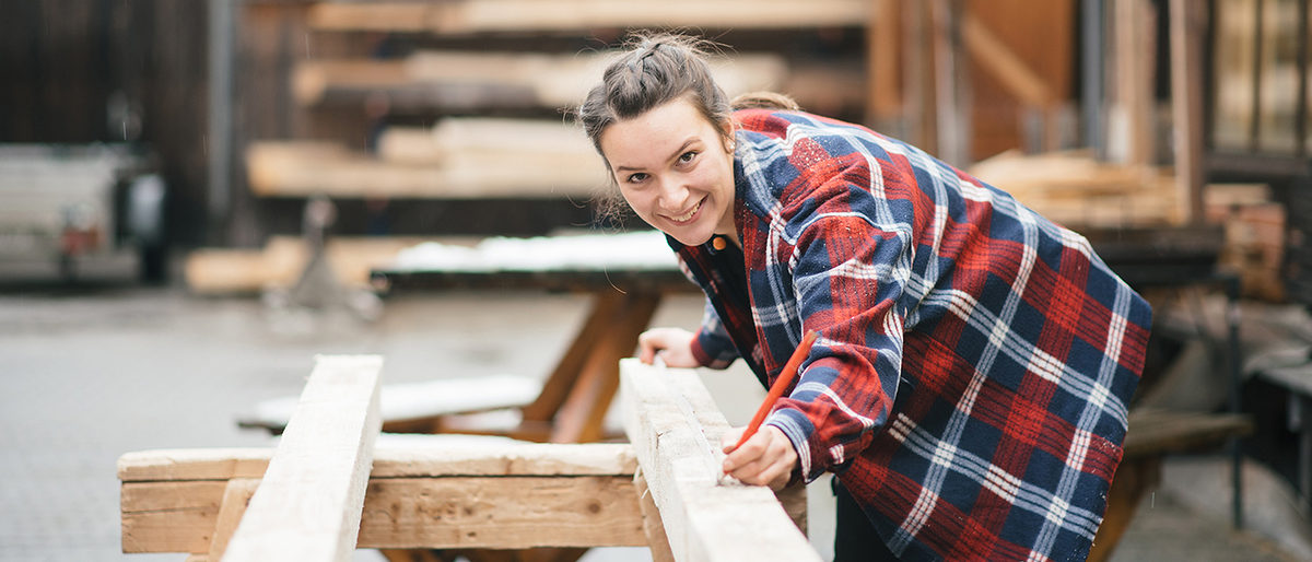 Teilzeitausbildung im Handwerk Junge Zimmerin markiert Holzbalken mit Bleistift und lächelt in die Kamera.