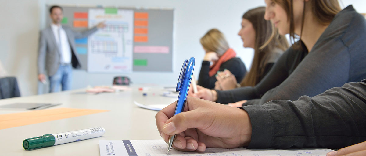 Berufsfachschule Schreibende Schüler, im Hintergrund an Tafel verschwommen der Lehrer.