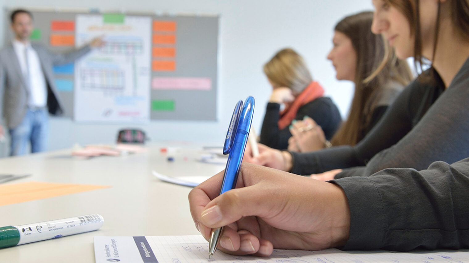 Schreibende Schüler, im Hintergrund an Tafel verschwommen der Lehrer.