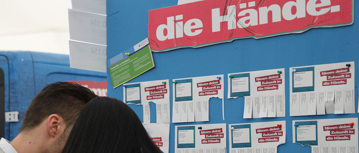 Jetzt Lehrstellen eintragen! Angeschnittene Köpfe von Junge und Mädchen von hinten vor Lehrstellenwand.