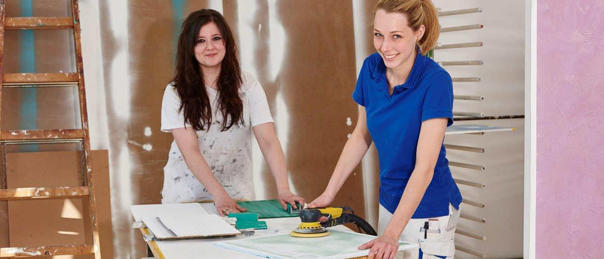 Zwei junge Maler und Lackiererinnen bei der Arbeit lächeln in die Kamera.