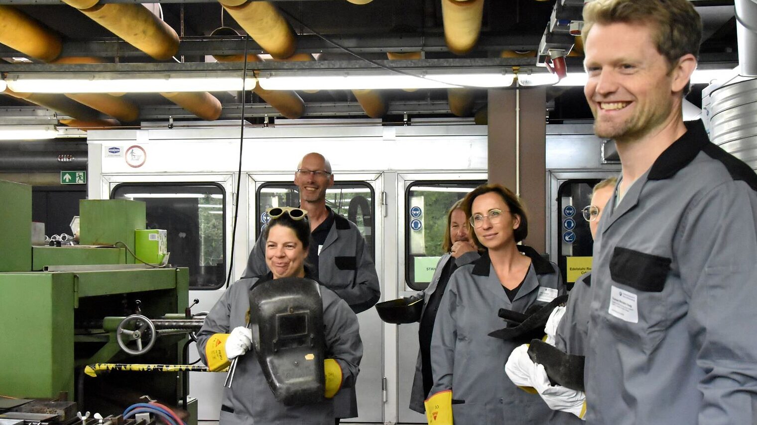 Sechs Lehrer stehen, zum Teil mit Schweißmasken in der Hand, in einem Raum mit Maschinen.