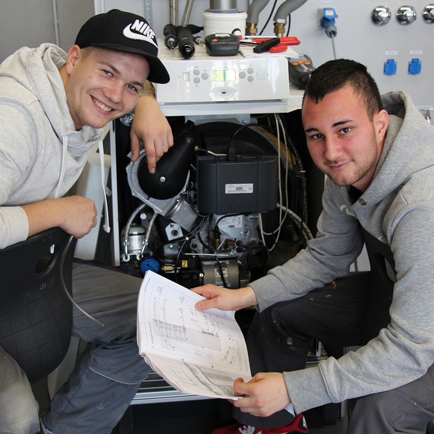 Zwei junge Anlagenmechaniker für Sanitär-, Heizungs- und Klimatechnik knien vor einem Gerät und lächeln in die Kamera.