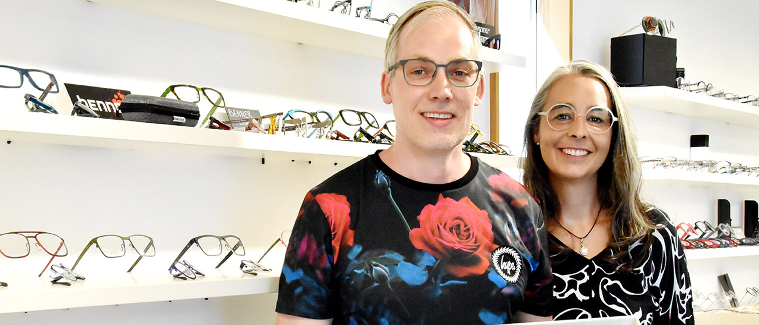 Augenoptiker- und Hörakustikermeister Daniel Benner steht mit seiner Frau vor einem Regal mit Brillen.