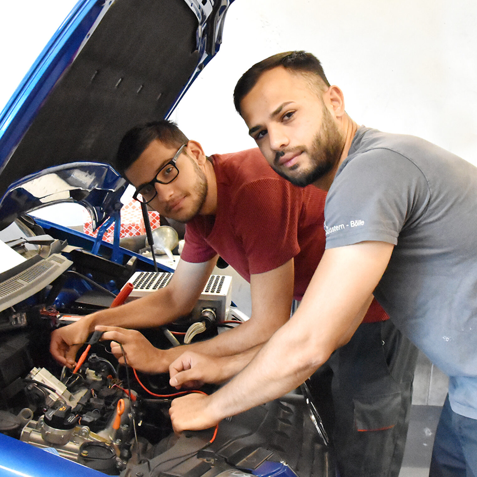 Zwei junge Kfz-Mechatroniker beugen sich unter geöffneter Motorhaube über den Motor eines Pkw, in der Bildungsakademie Singen.