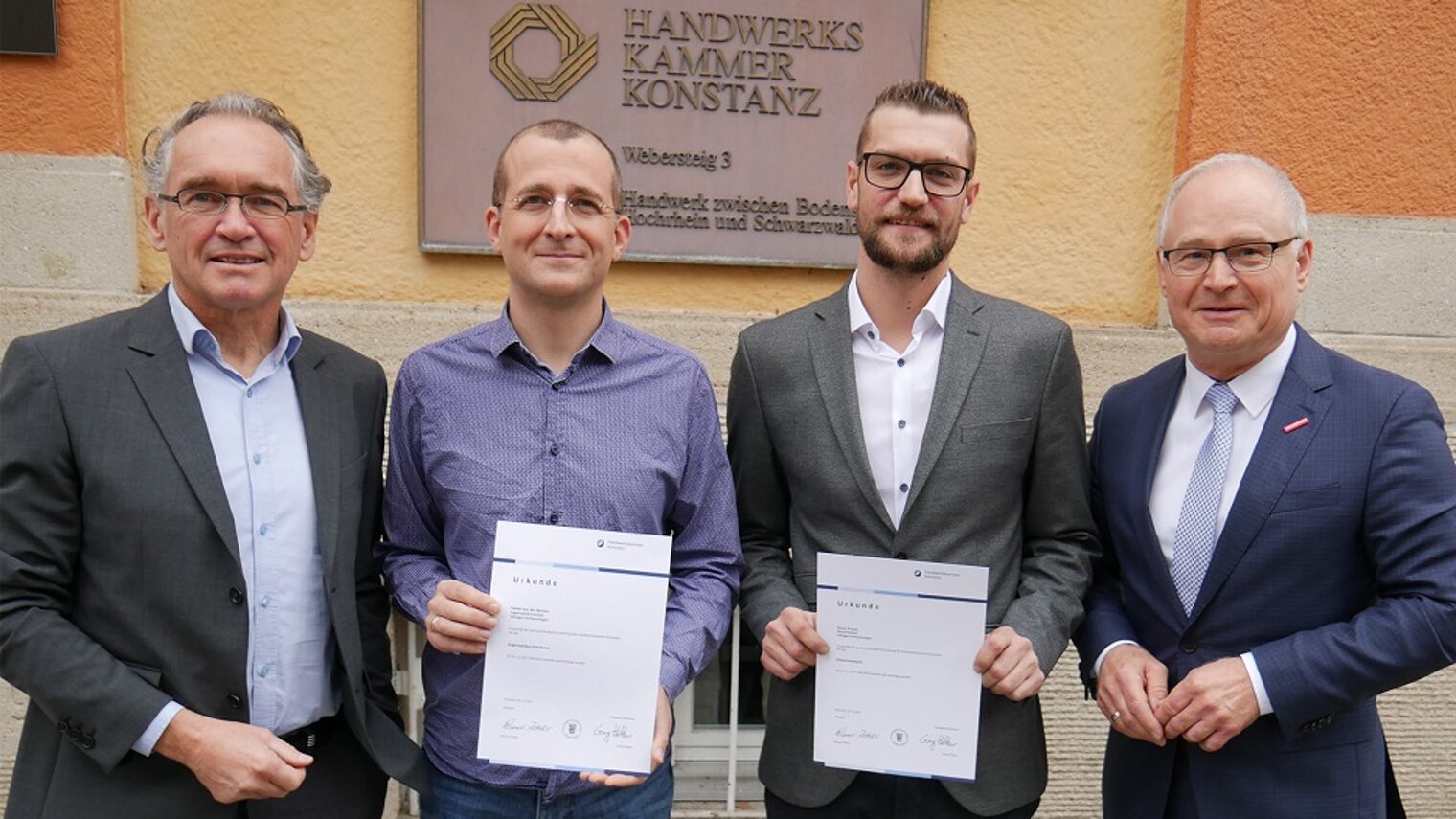 Vier Männer schauen zur Kamera. Die beiden mittleren Herren zeigen Urkunden.