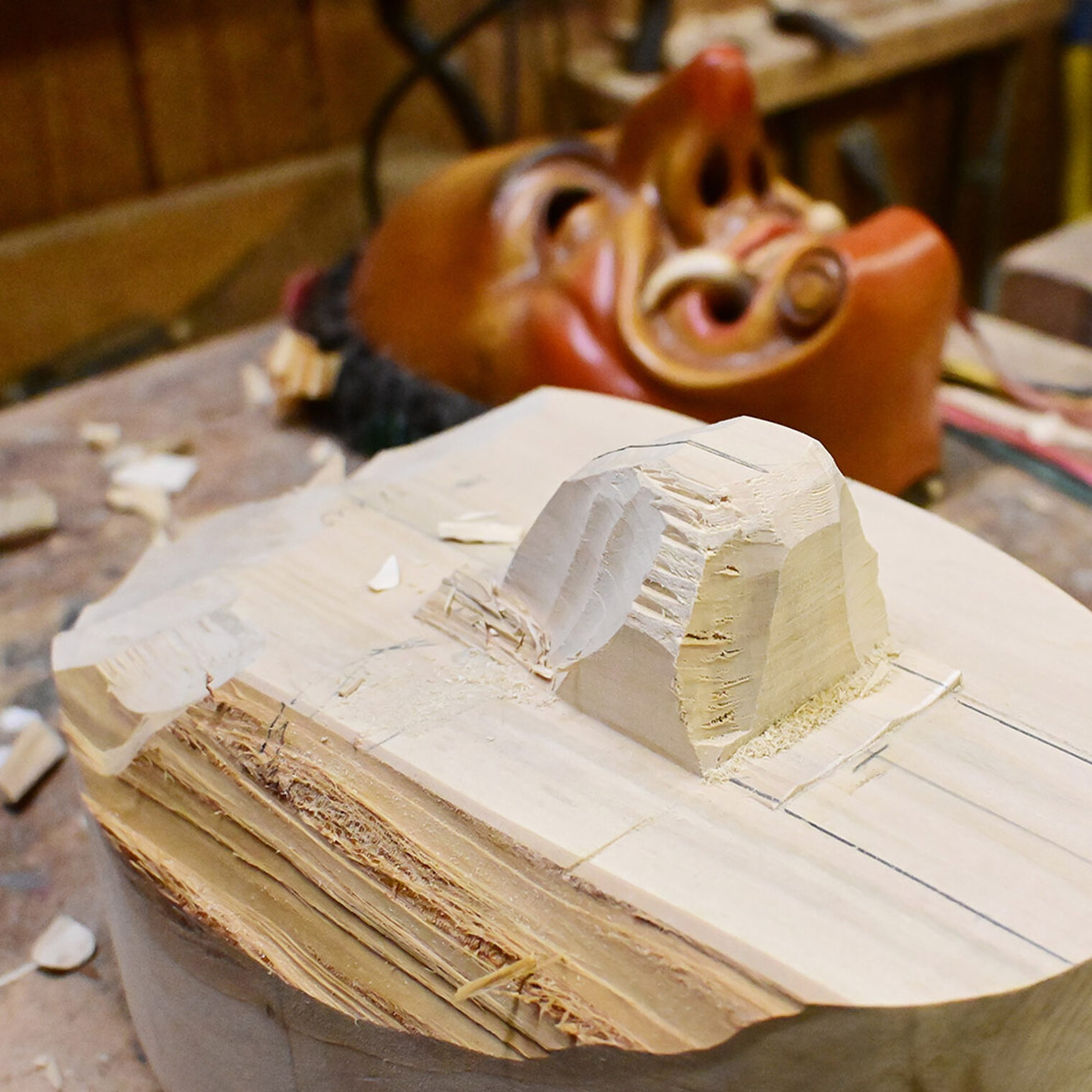 Im Vordergrund ein nur leicht bearbeiteter Holzklotz, im Hintergrund eine fertige Fasnachts-Larve.