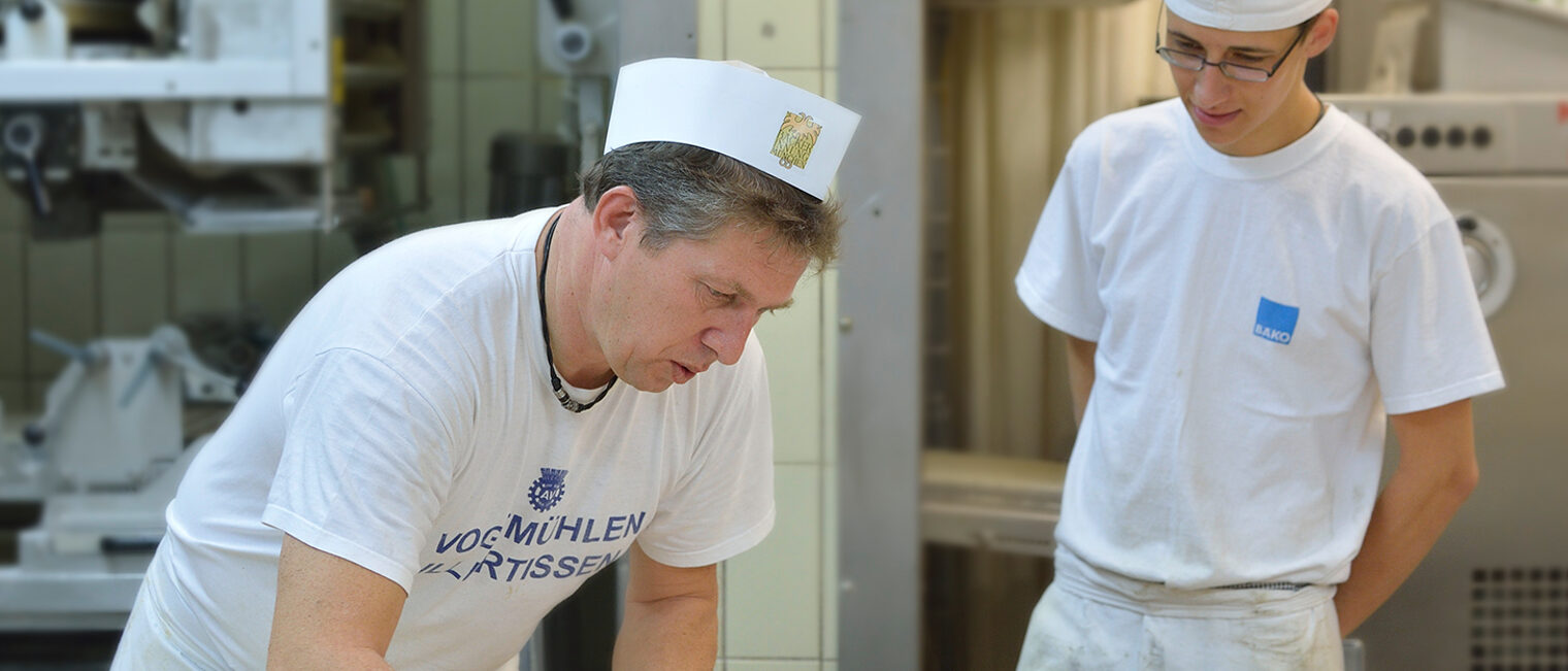 Bäcker knetet Teig in der Backstube, Lehrling oder Schüler schaut zu.
