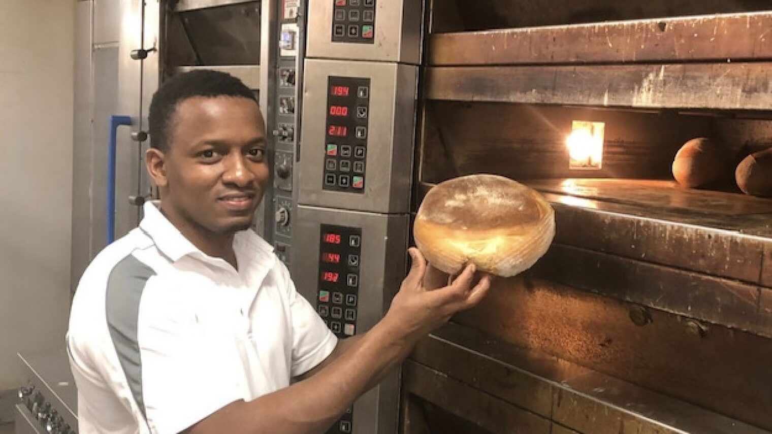 Junger Mann mit einem Brot in der Hand steht vor großem Bäckerofen