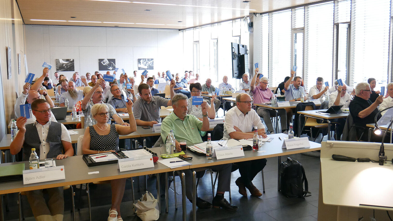 Vollversammlungsmitglieder sitzen an Tischen und heben Stimmkarten.