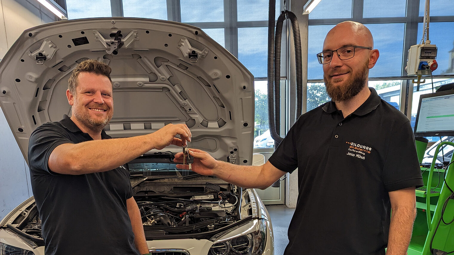 Der alte Kfz-Lehrmeister Markus Maurer übergibt einen Autoschlüssel an seinen Nachfolger Jonas Münch, vor geöffneter Motorhaube.