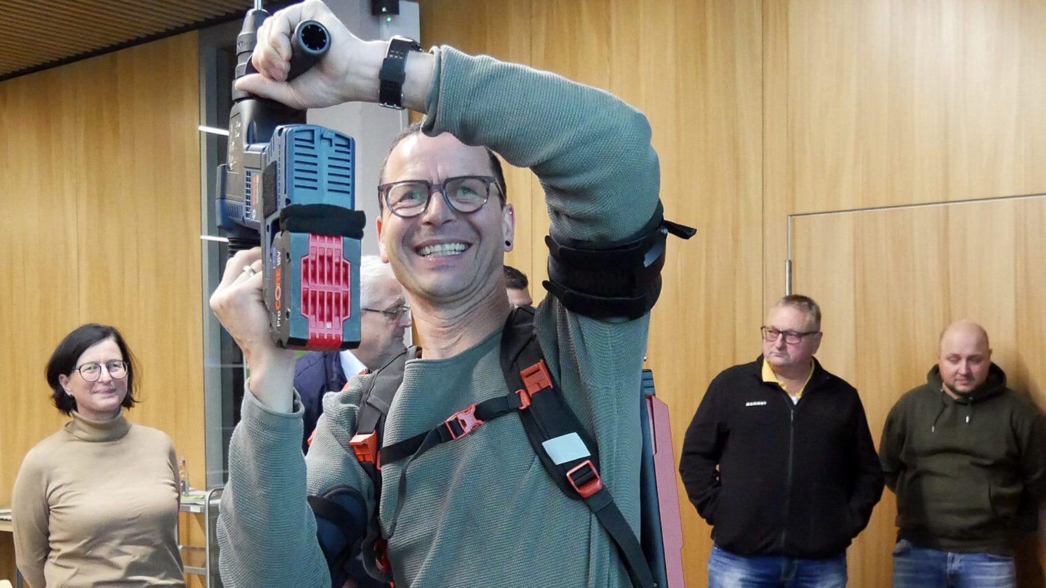 Malermeister Joachim Würstl testet mit erhobener Bohrmaschine ein Exoskelett, das er wie einen Rucksack trägt.