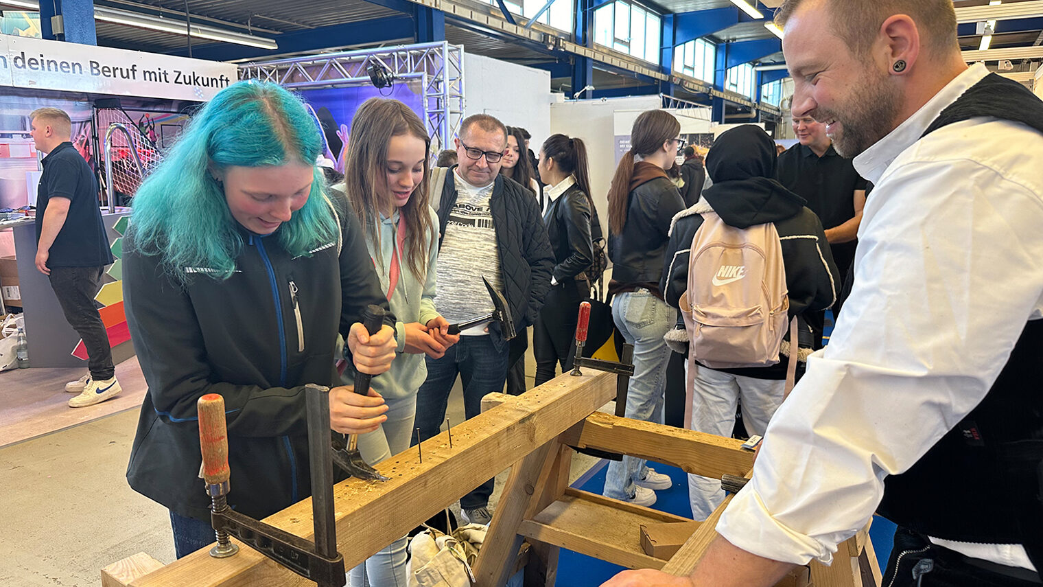 Auf der Messe Jobs für Future in Villingen schlagen zwei Mädchen Nägel in einen Holzbalken. Ein Zimmerer leitet sie an.