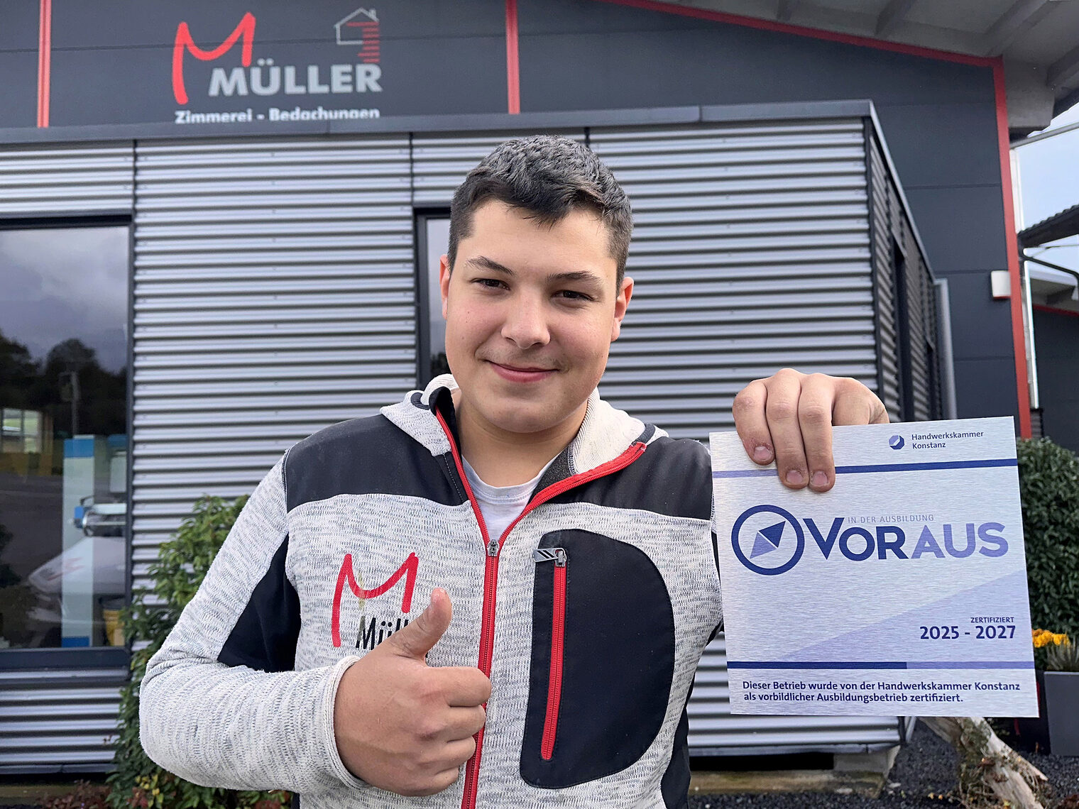 Azubi Felix Mess mit VORAUS-Schild in der einen Hand und Daumen hoch mit der anderen vor dem Firmengebäude von Holzbau Müller.