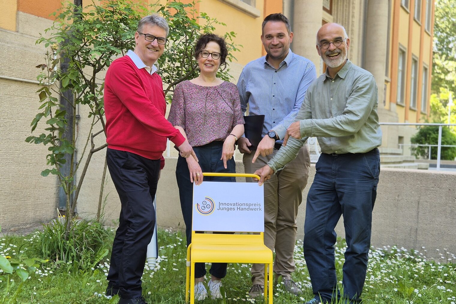 Vier Personen hinter einem gelben Stuhl weisen mit ihren Händen auf ein Schild zum Innovationspreis
