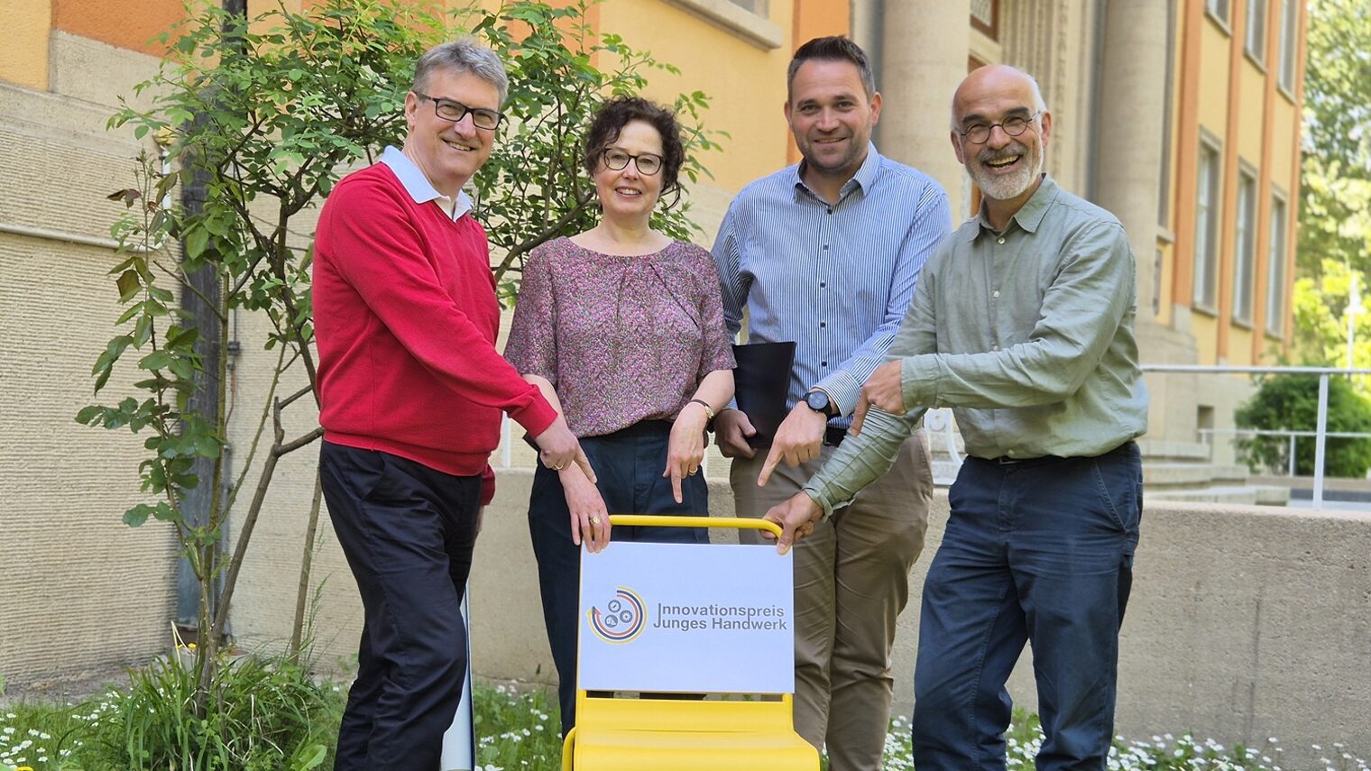 Vier Personen hinter einem gelben Stuhl weisen mit ihren Händen auf ein Schild zum Innovationspreis