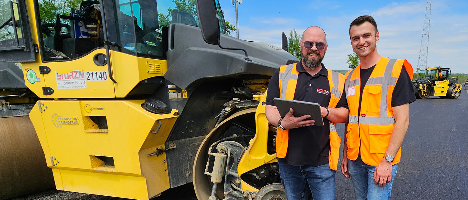 Zwei Straßenbauer mit Tablet vor einer großen Walzmaschine.
