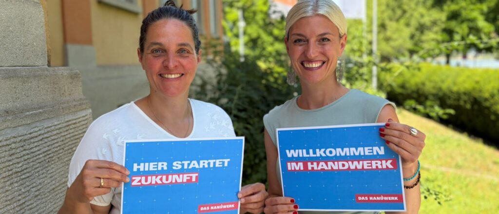 Zwei Frauen stehen im Freien, lächeln in die Kamera und halten jeweils ein Schild hoch, auf denen steht "Hier startet Zukunft" und "Willkommen im Handwerk".