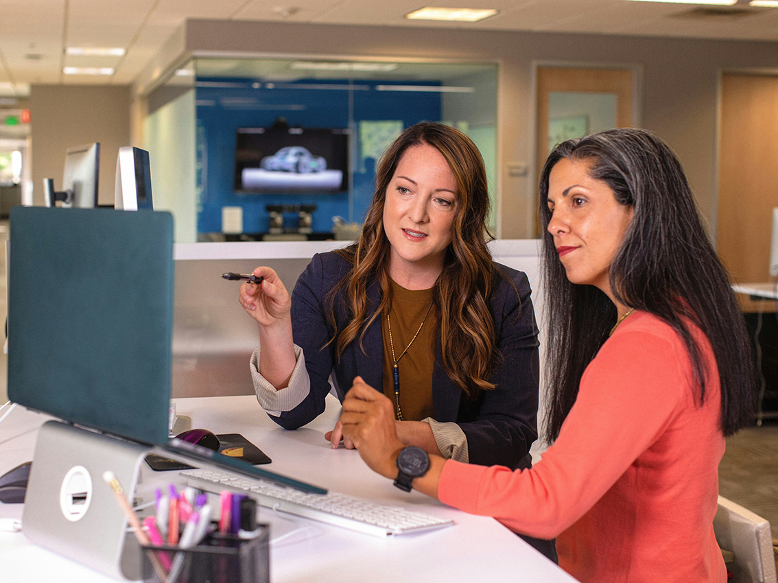 Einer Frau erklärt einer anderen etwas am PC-Bildschirm.