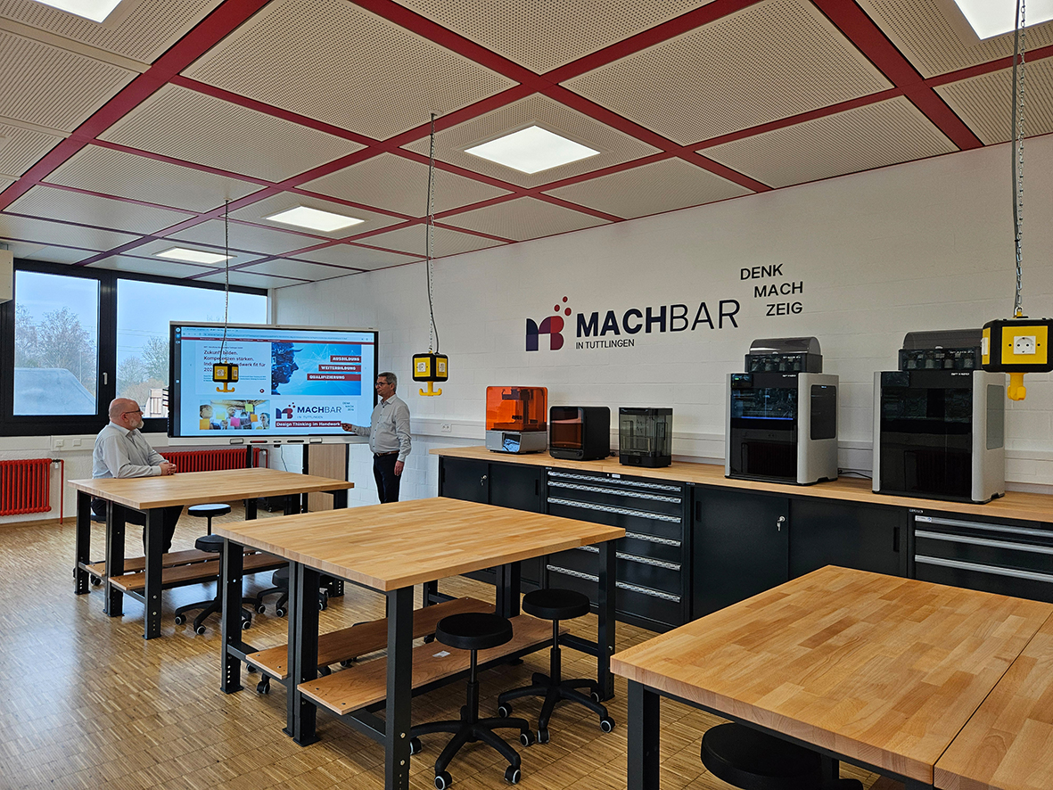 Kreativraum in Tuttlingen mit Holztischen, zwei Personen vor Smartboard und diversen 3D-Druckern auf einem Sideboard vor einer Wand.