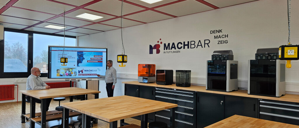 Kreativraum in Tuttlingen mit Holztischen, zwei Personen vor Smartboard und diversen 3D-Druckern auf einem Sideboard vor einer Wand.