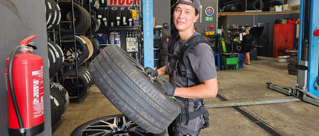 Mann in Kfz-Werkstatt hebt einen Autoreifen an. Er trägt ein Exoskelett.