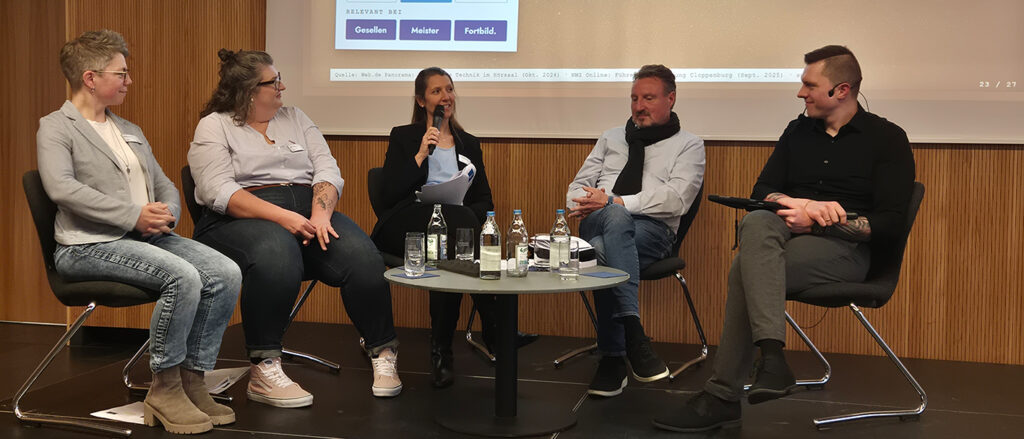 Expertenrunde aus drei Frauen und zwei Männern sitzt auf Podium um einen kleinen runden Tisch. Im Hintergrund sieht man einen Teil einer Power-Point-Präsentation.