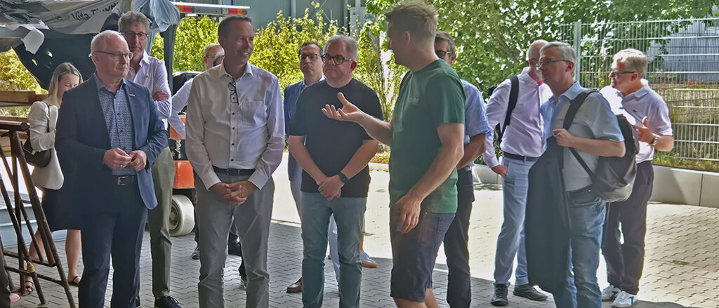 Eine Delegation mit 13 Männern steht vor einer Halle der Schreinerei Huber. Firmenchef Huber spricht zu Staatssekretär Dörflinger.