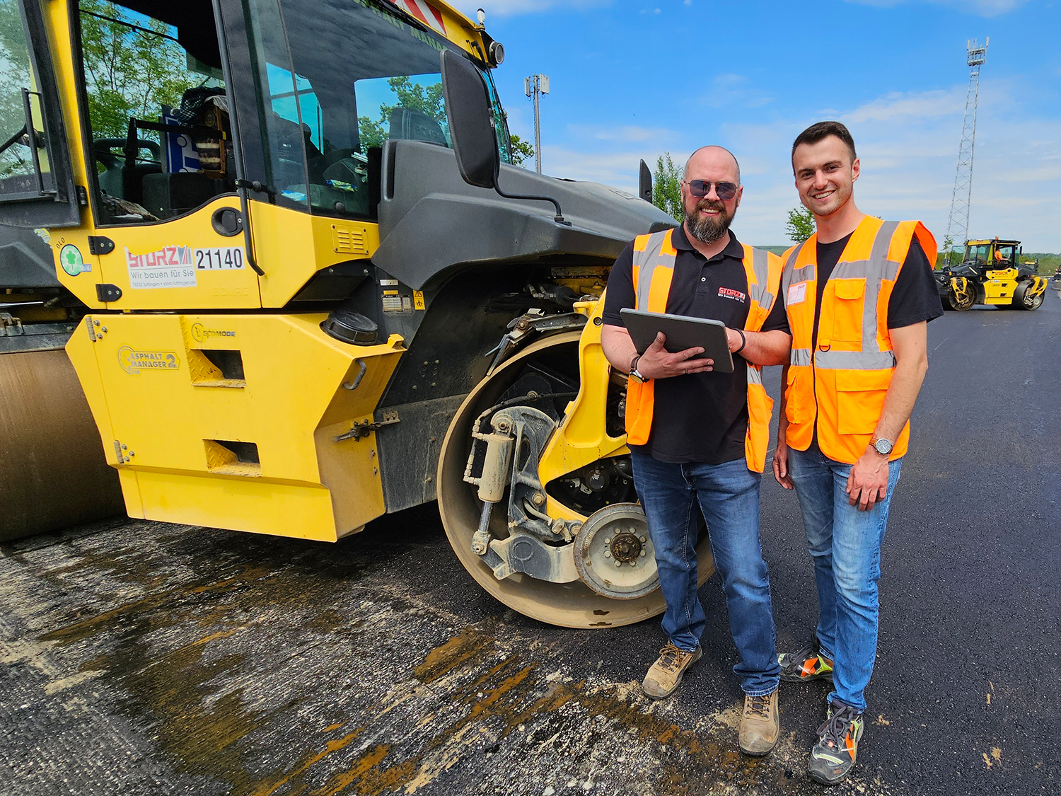 Zwei Straßenbauer mit Tablet vor einer großen Walzmaschine.