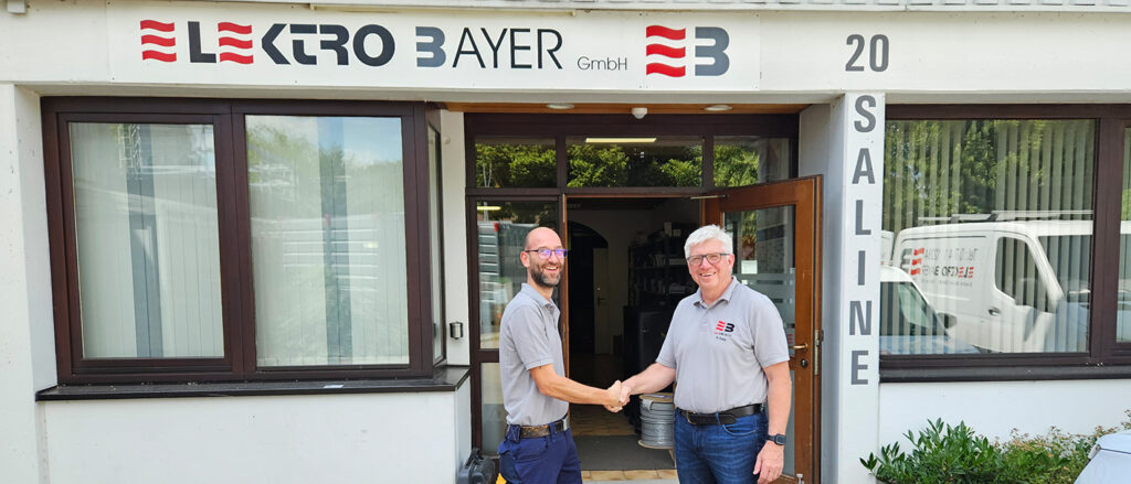 Ulrich Kammerer-Romano und Ralf Rapp geben sich vor dem Firmengebäude der Elektro Bayer Gmbh die Hand und lächeln in die Kamera.