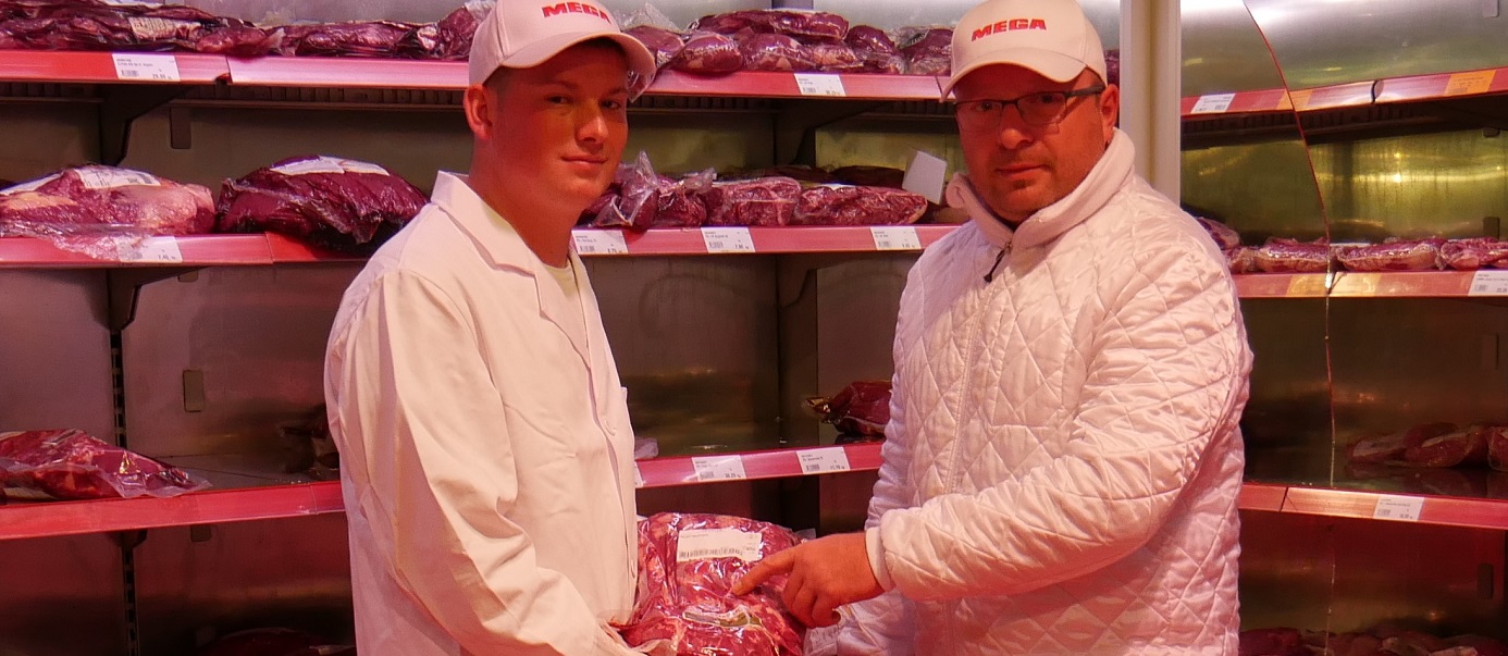 Ausbilder und ehemaliger Azubi zwischen Regalen voller Fleisch.