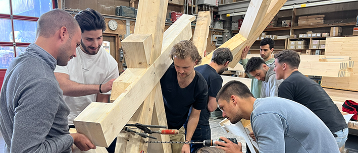 Mehrere Studenten umringen einen Fachwerkknoten in einer Zimmerei-Werkstatt.