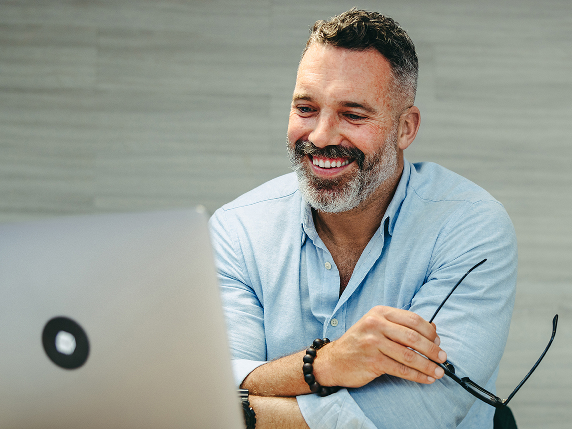 Mann mit graumeliertem Bart hält seine Brille in der Hand und lächelt in einen geöffneten Laptop.