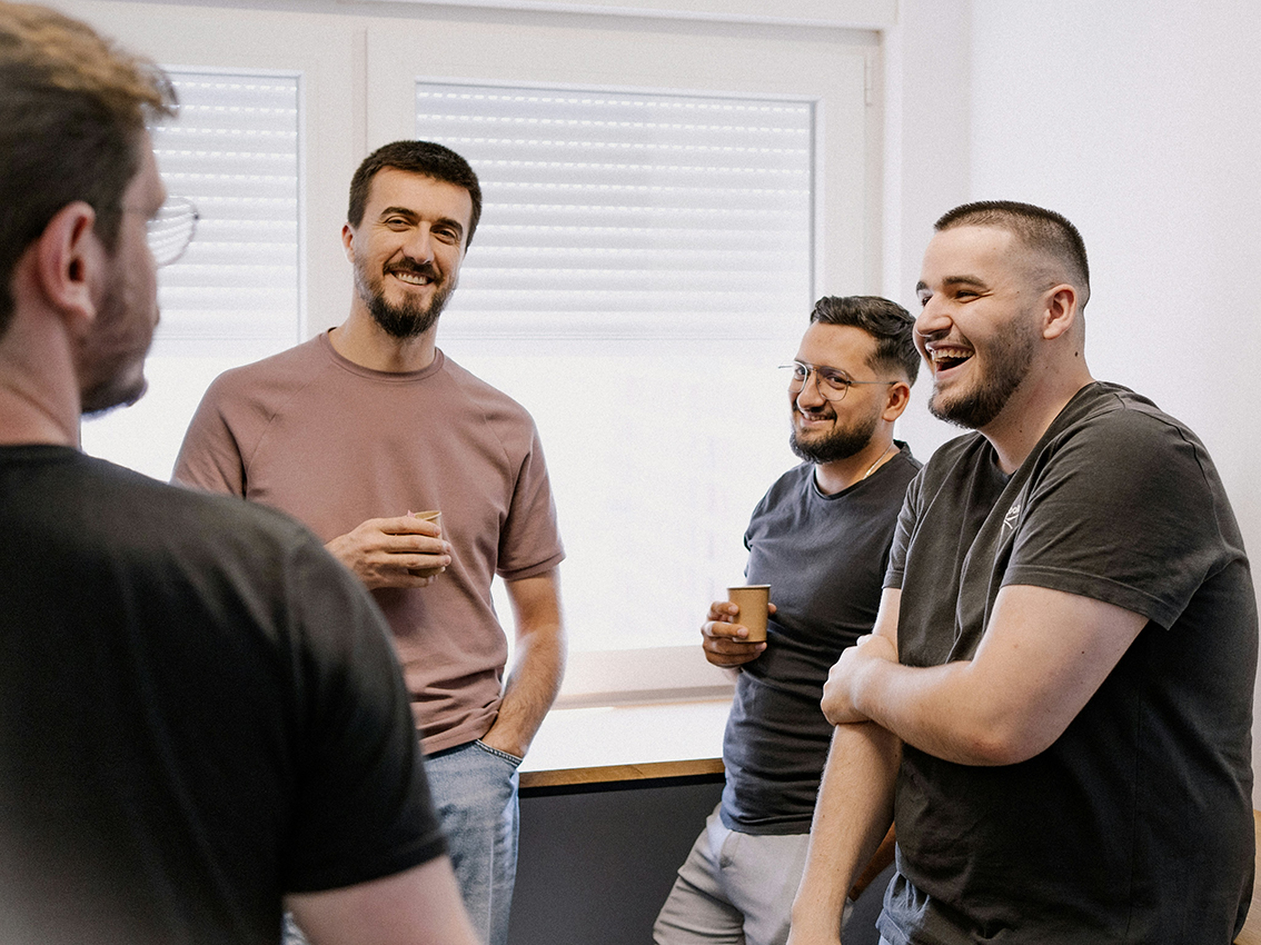 Vier mittelalte Männer stehen mit Kaffeebechern in der Hand beisammen und lachen.